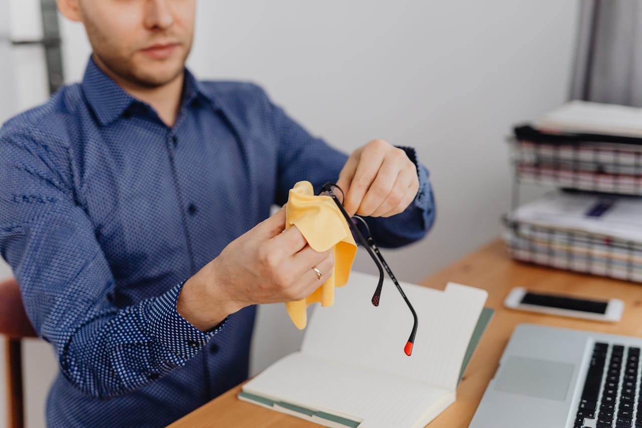 our-story Young man in blue shirt cleaning eyeglasses with cloth at his desk.